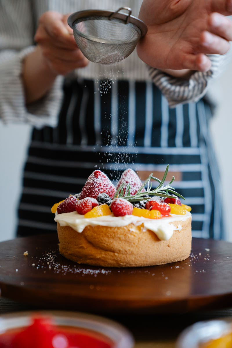 Closeup of a chef sprinkling powdered sugar on a cream-topped cake with strawberries.