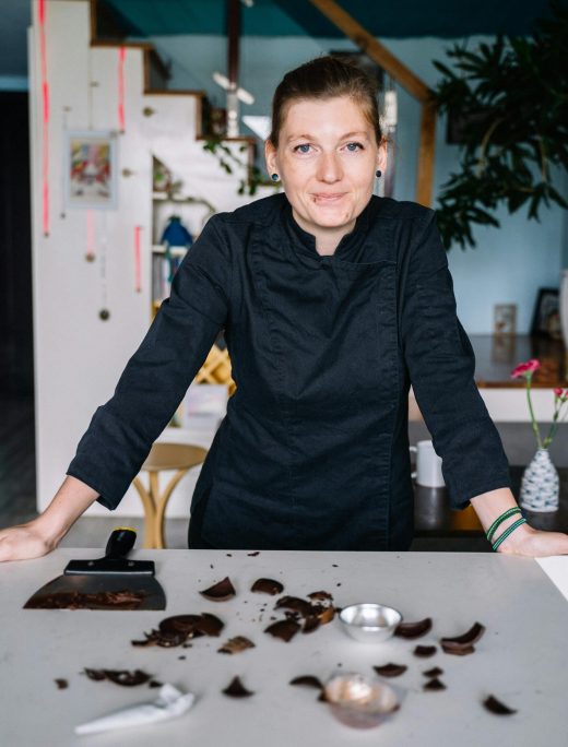 Smiling chef in black uniform with chocolate pieces and tools on a kitchen counter.